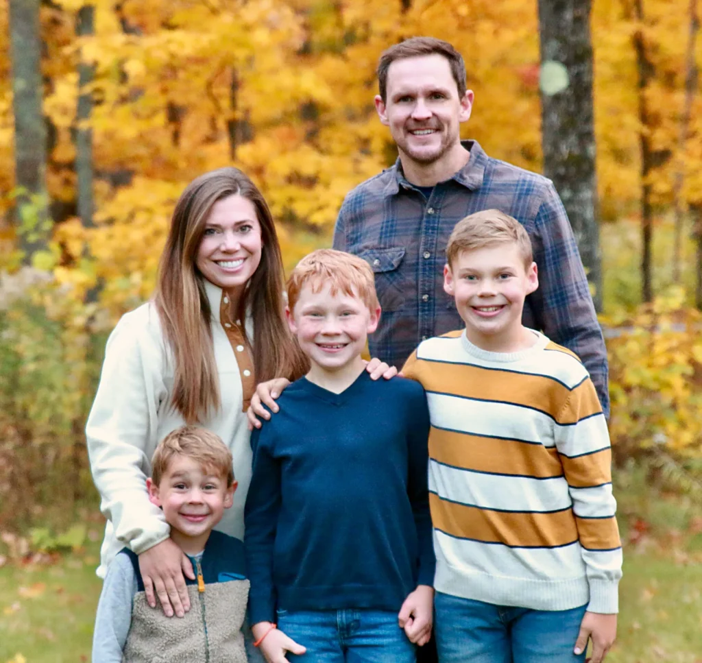 A family of five stands together outdoors in front of autumn leaves, reminiscent of Esko, MN's charm. The parents smile behind their three young boys, who are dressed in casual sweaters and jeans. The vibrant fall foliage provides a colorful backdrop, like the rich hues of pure maple syrup.